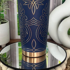 May include: A navy blue tumbler with gold accents and an intricate gold design. The tumbler has a gold rim and base. The design features floral and geometric patterns. The tumbler is displayed on a mirrored surface.