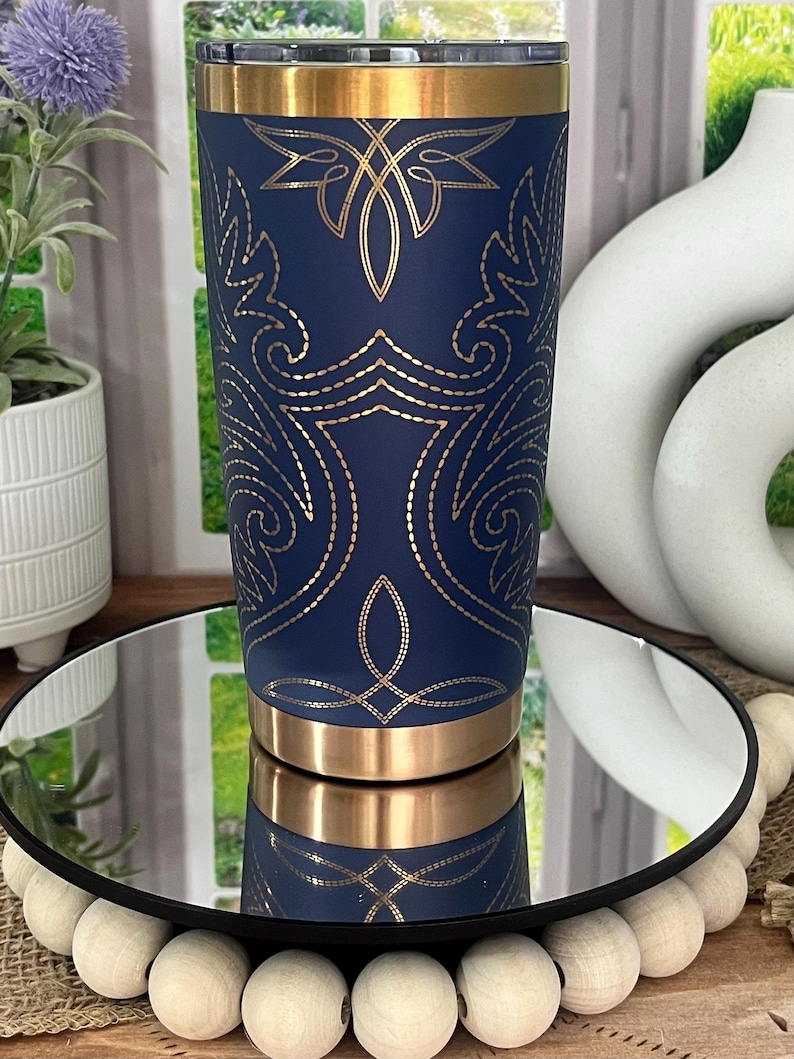 May include: A navy blue tumbler with gold accents and an intricate gold design. The tumbler has a gold rim and base. The design features floral and geometric patterns. The tumbler is displayed on a mirrored surface.
