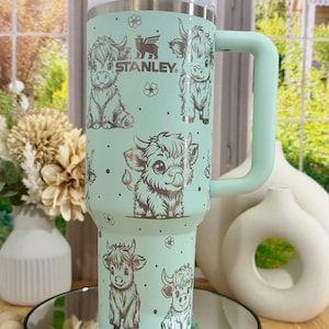 May include: A mint green Stanley tumbler with a white lid and handle. The tumbler is decorated with cartoon illustrations of Highland cows and small flowers. The Stanley logo is visible on the front.