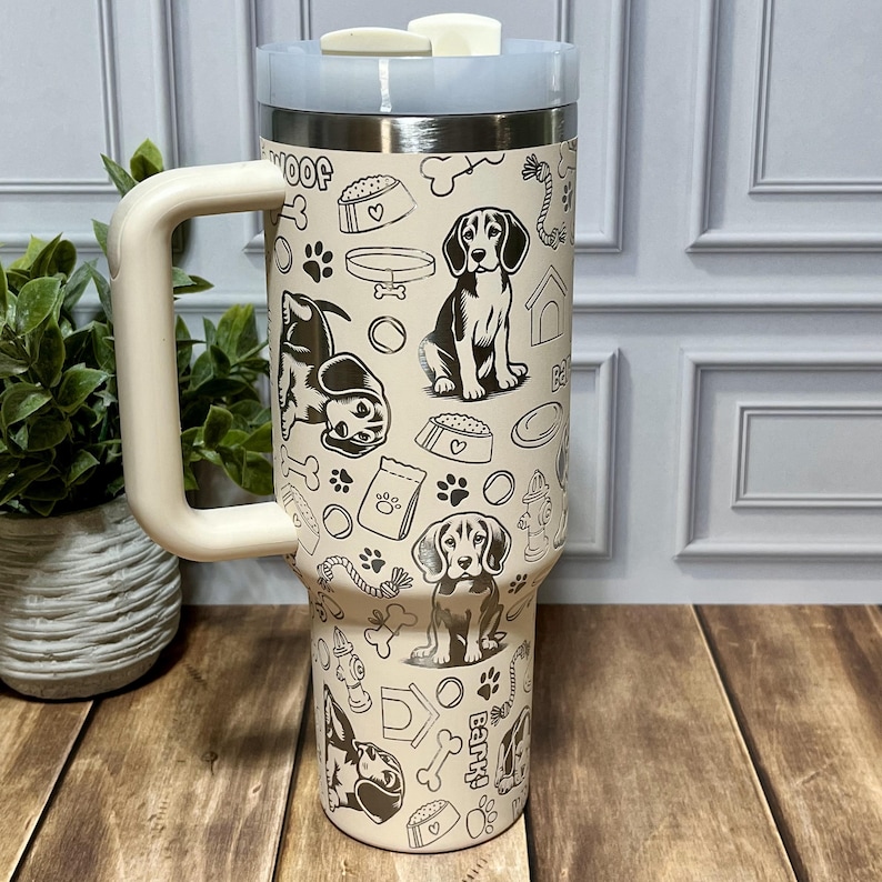 May include: A beige travel mug with a light blue lid and handle, featuring a repeating black and white dog-themed pattern. The pattern includes dog illustrations, paw prints, food bowls, and bones. The mug has a large handle.