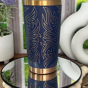 May include: A navy blue tumbler with gold accents and a gold floral design. The stainless steel tumbler is cylindrical and has a gold band at the top and bottom. The tumbler is displayed on a mirrored surface.