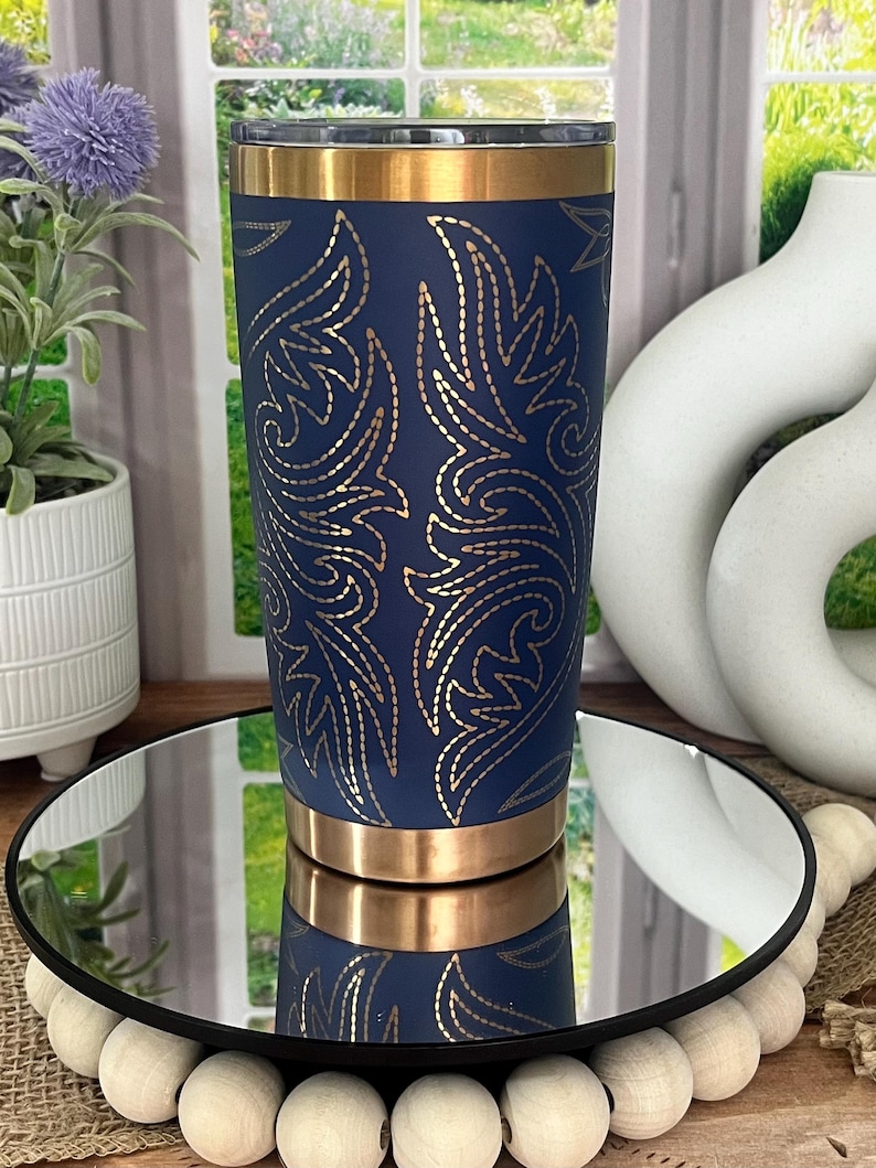 May include: A navy blue tumbler with gold accents and a gold floral design. The stainless steel tumbler is cylindrical and has a gold band at the top and bottom. The tumbler is displayed on a mirrored surface.