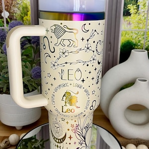 May include: Cream-colored travel mug with a handle, featuring a Leo zodiac design. The design includes a lion, constellations, and the word "Leo." The mug has a white lid and a rainbow-colored band near the top.