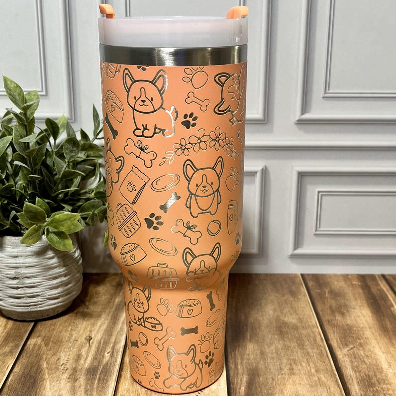 K&ouml;nnte beinhalten: Ein Edelstahlbecher in Pfirsichfarbe, verziert mit einem sich wiederholenden Muster aus Hundeillustrationen, Pfotenabdr&uuml;cken und hundebezogenen Symbolen. Der Becher hat einen wei&szlig;en Deckel und einen orangefarbenen Griff. Ein dekorativer Artikel f&uuml;r Hundeliebhaber.