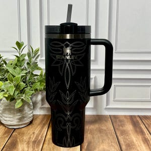 May include: Black Stanley tumbler with a handle and a straw. The tumbler features a decorative silver design and the Stanley logo. The tumbler is on a wooden surface with a plant in the background.