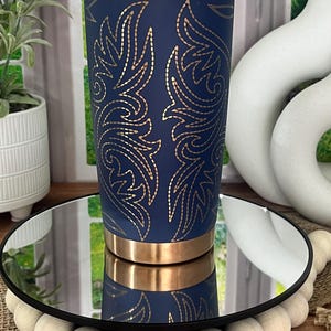 May include: A navy blue insulated tumbler with a gold floral design. The tumbler has a gold rim and base. The design is symmetrical and covers the majority of the tumbler's surface. The tumbler is displayed on a mirrored surface.
