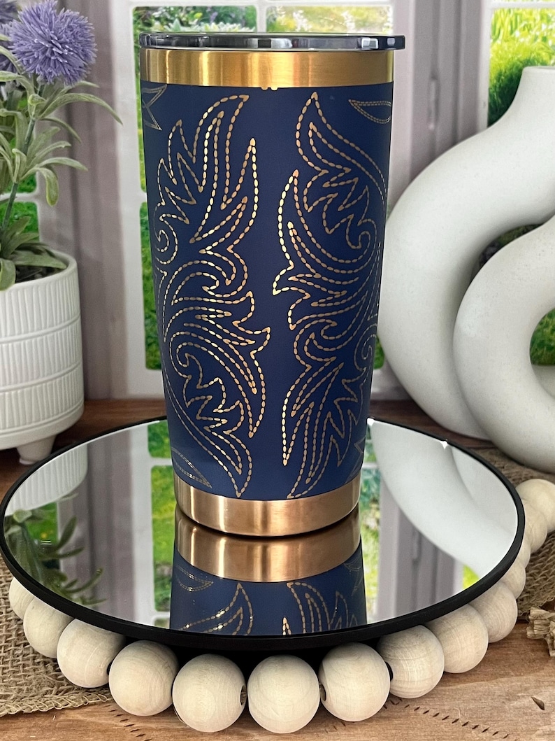 May include: A navy blue insulated tumbler with a gold floral design. The tumbler has a gold rim and base. The design is symmetrical and covers the majority of the tumbler's surface. The tumbler is displayed on a mirrored surface.