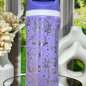 May include: A purple insulated tumbler with a white and purple flip-top lid. The tumbler is decorated with a gold pattern of cartoon Highland cows and gold dots. The tumbler is sitting on a mirrored surface.