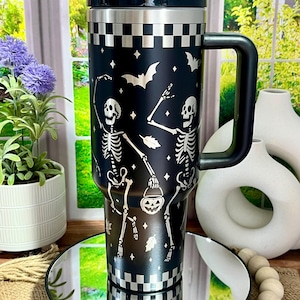 May include: Black travel mug with a handle, featuring a white skeleton design, bats, and a jack-o'-lantern. The mug has a checkered pattern at the top and bottom. The lid and handle are black.