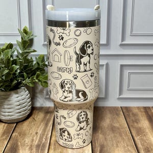 May include: A beige stainless steel tumbler with a light blue lid, featuring a black and white dog-themed pattern. The design includes dog illustrations, paw prints, bones, and the word "Bark!" and "Woof!"
