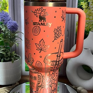 May include: An orange Stanley tumbler with a handle, featuring gold leaf and pumpkin spice designs. The tumbler has a clear lid and a straw. The Stanley logo is visible. The tumbler is designed for cold beverages.