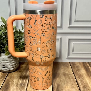 K&ouml;nnte beinhalten: Ein orangefarbener isolierter Becher mit Griff, der mit einem silbernen Muster aus Corgi-Hunden, Knochen und Pfotenabdr&uuml;cken verziert ist. Der Becher hat einen wei&szlig;en Deckel und einen orangefarbenen Deckel. Der Griff und der Deckel haben den gleichen Orangeton wie der Becher.