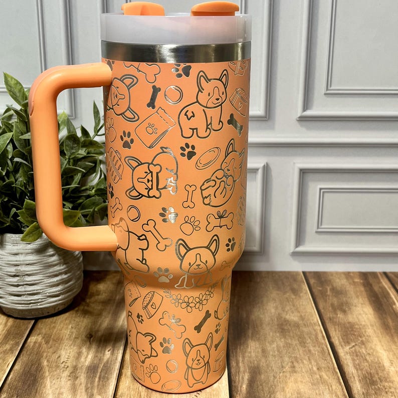 K&ouml;nnte beinhalten: Ein orangefarbener isolierter Becher mit Griff, der mit einem silbernen Muster aus Corgi-Hunden, Knochen und Pfotenabdr&uuml;cken verziert ist. Der Becher hat einen wei&szlig;en Deckel und einen orangefarbenen Deckel. Der Griff und der Deckel haben den gleichen Orangeton wie der Becher.