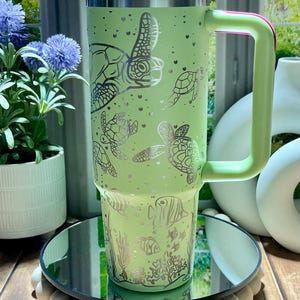 May include: A light green insulated tumbler with a white lid and a green handle. The tumbler features a silver design of sea turtles, fish, and coral. The handle has a pink accent. The tumbler is approximately 10 inches tall.