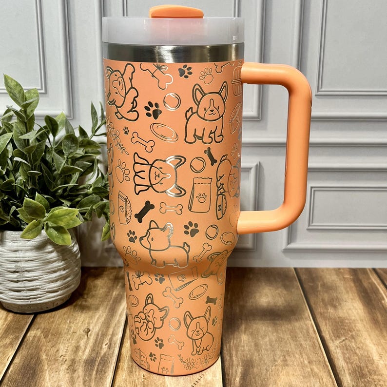K&ouml;nnte beinhalten: Ein pfirsichfarbener Reisebecher mit einem silbernen Hunde-Muster. Das Design umfasst Hundeillustrationen, Pfotenabdr&uuml;cke und Knochen. Er hat einen passenden pfirsichfarbenen Griff und Deckel sowie eine transparente Kunststoffoberseite.