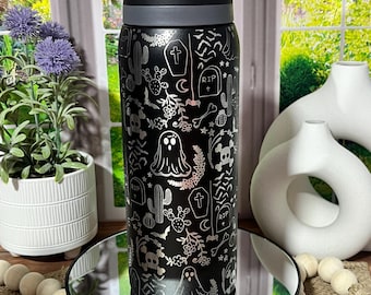 May include: Black water bottle with a black lid and a grey band. The bottle features a silver Halloween-themed pattern of ghosts, skulls, cacti, and tombstones. The bottle is on a mirrored surface.