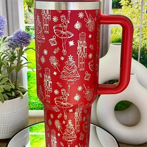 May include: A red insulated tumbler with a matching handle and lid. The tumbler is decorated with a silver pattern of Christmas trees, ballerinas, and nutcrackers. The tumbler is designed to hold approximately 30 ounces.