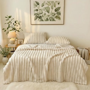 Puede incluir: Una cama acogedora cubierta con una manta crema con costuras verticales y dos almohadas a juego. Una impresión botánica enmarcada cuelga sobre la cama. Una pequeña mesa auxiliar con una lámpara y vegetación está a la izquierda.