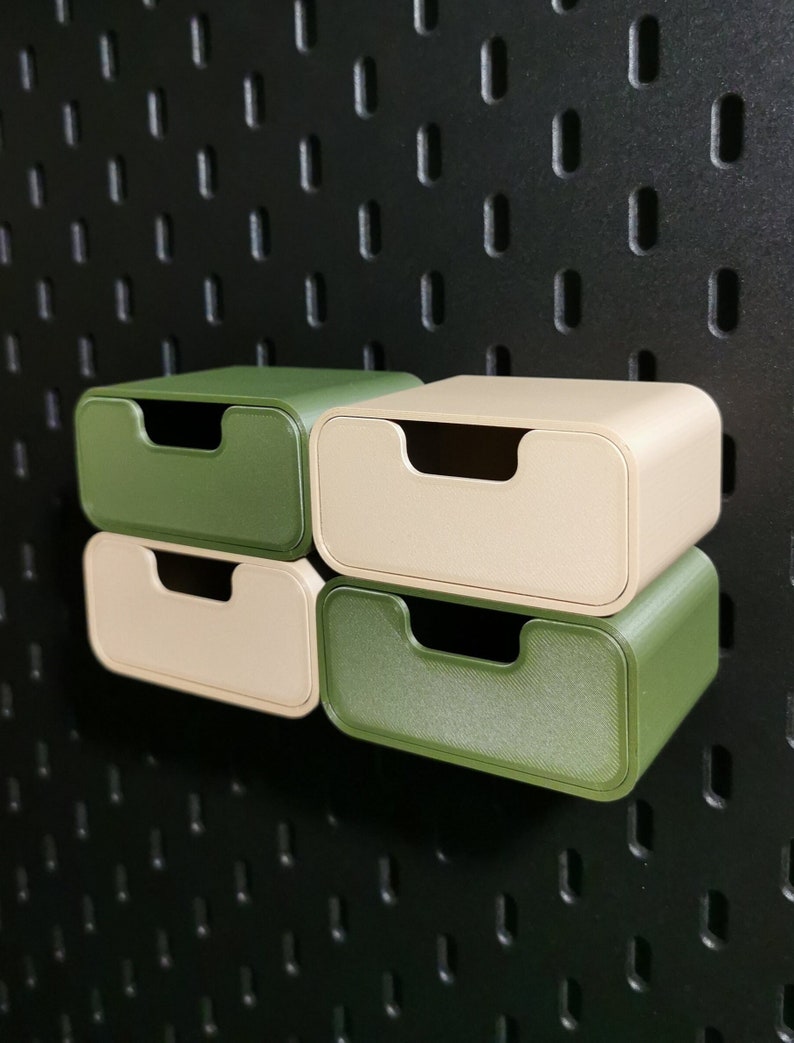 Puede incluir: Cuatro peque&ntilde;os contenedores de almacenamiento rectangulares en verde oliva y beige apilados contra una pared perforada negra. Cada contenedor tiene una parte superior curva con un asa empotrada. Los contenedores son de material texturizado.