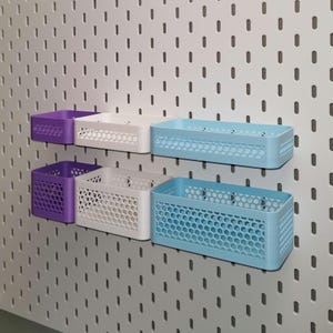 Op de afbeelding: Een set van zes rechthoekige opbergmanden in paars, wit en lichtblauw, gemonteerd op een wit pegboard. De manden hebben een honingraatpatroon en zijn ontworpen voor organisatie en display.