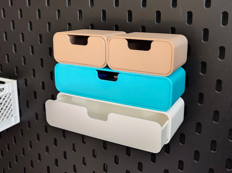 Puede incluir: Cuatro cajones de almacenamiento coloridos en beige, turquesa y blanco est&aacute;n montados en una pared perforada negra. Los cajones son rectangulares con bordes redondeados y un asa empotrada. Los dos cajones superiores son beige, el del medio es turquesa y el inferior es blanco.