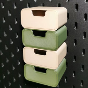 Puede incluir: Cuatro peque&ntilde;os cajones de almacenamiento rectangulares en beige y verde apilados contra una pared perforada negra. Cada caj&oacute;n tiene un recorte de asa curvado. Los cajones son de material texturizado.