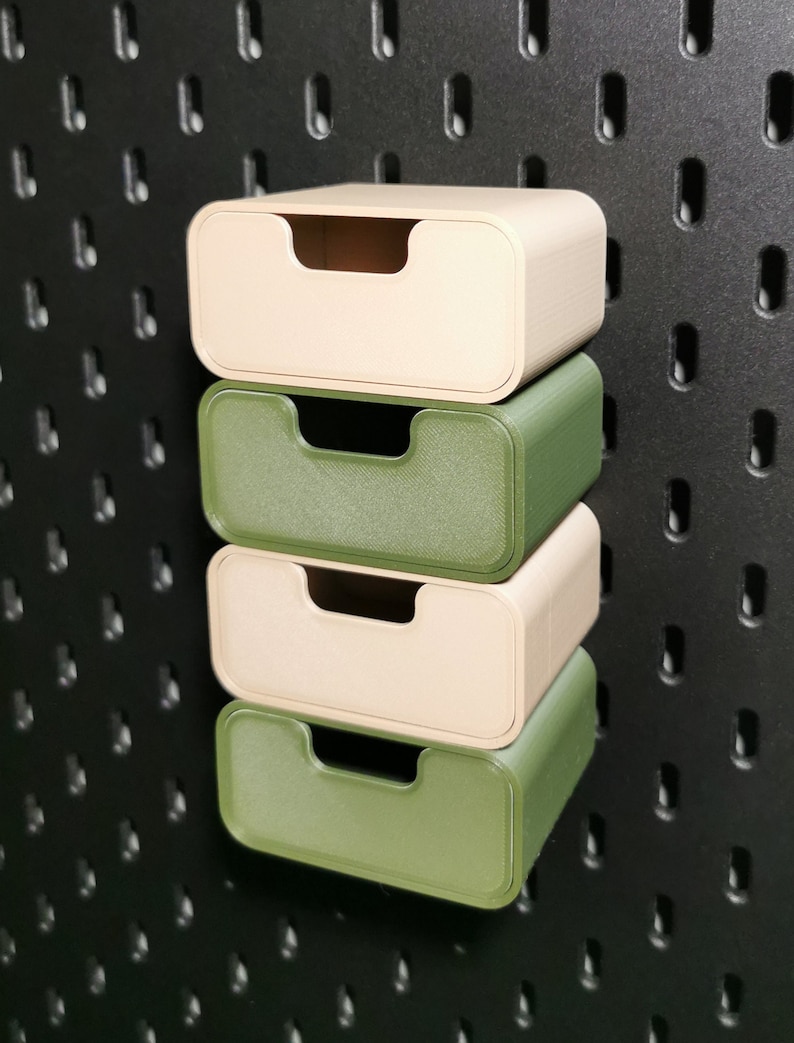 Puede incluir: Cuatro peque&ntilde;os cajones de almacenamiento rectangulares en beige y verde apilados contra una pared perforada negra. Cada caj&oacute;n tiene un recorte de asa curvado. Los cajones son de material texturizado.