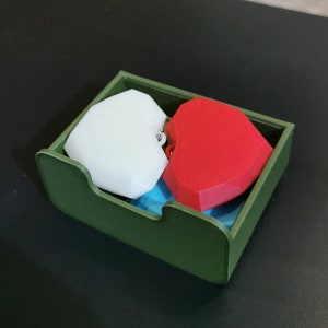 Puede incluir: Un contenedor rectangular verde que contiene dos objetos en forma de coraz&oacute;n. Un coraz&oacute;n es blanco y el otro es rojo. El contenedor tiene un frente curvo y est&aacute; sobre una superficie oscura.