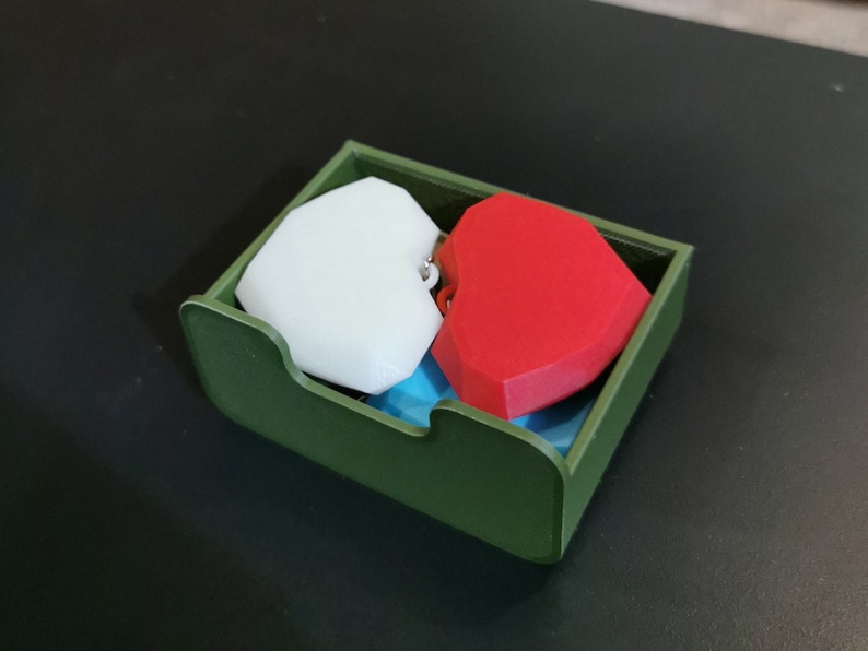 Puede incluir: Un contenedor rectangular verde que contiene dos objetos en forma de coraz&oacute;n. Un coraz&oacute;n es blanco y el otro es rojo. El contenedor tiene un frente curvo y est&aacute; sobre una superficie oscura.