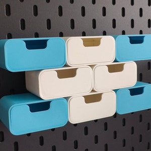 Puede incluir: Un conjunto de peque&ntilde;os contenedores de almacenamiento rectangulares en azul claro y blanco roto. Los contenedores tienen un dise&ntilde;o redondeado con un asa empotrada. Est&aacute;n dispuestos sobre una panel perforado gris oscuro, ofreciendo una soluci&oacute;n de almacenamiento moderna.