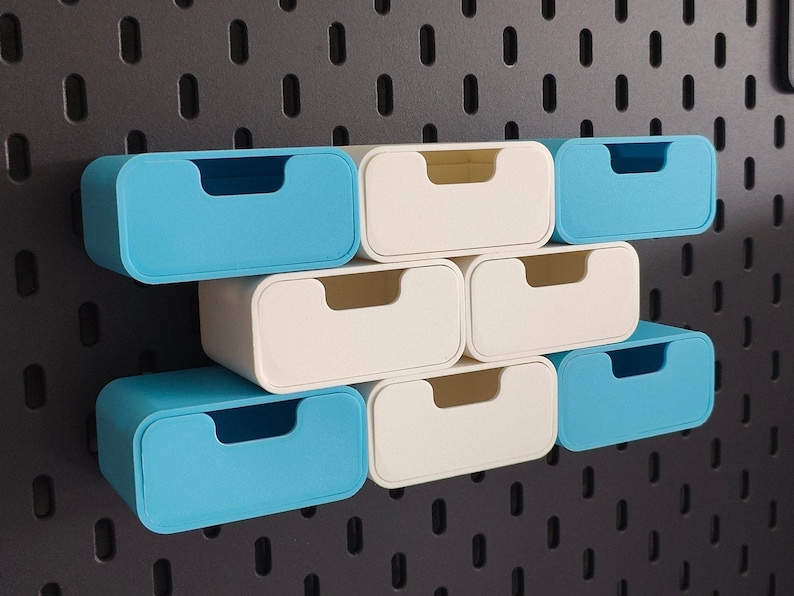 Puede incluir: Un conjunto de peque&ntilde;os contenedores de almacenamiento rectangulares en azul claro y blanco roto. Los contenedores tienen un dise&ntilde;o redondeado con un asa empotrada. Est&aacute;n dispuestos sobre una panel perforado gris oscuro, ofreciendo una soluci&oacute;n de almacenamiento moderna.