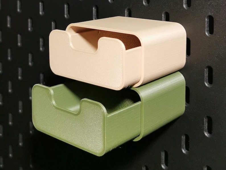 Puede incluir: Dos contenedores de almacenamiento impresos en 3D, uno beige y otro verde oliva, est&aacute;n apilados contra un panel perforado negro. Cada contenedor tiene un dise&ntilde;o curvo con una abertura frontal empotrada. Los contenedores est&aacute;n dise&ntilde;ados para montaje en pared.