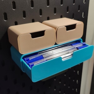 Puede incluir: Un conjunto de tres contenedores de almacenamiento en beige y turquesa. Los dos contenedores beige superiores tienen un dise&ntilde;o de caj&oacute;n. El contenedor turquesa inferior contiene bol&iacute;grafos azules. Montados en una pared perforada negra.
