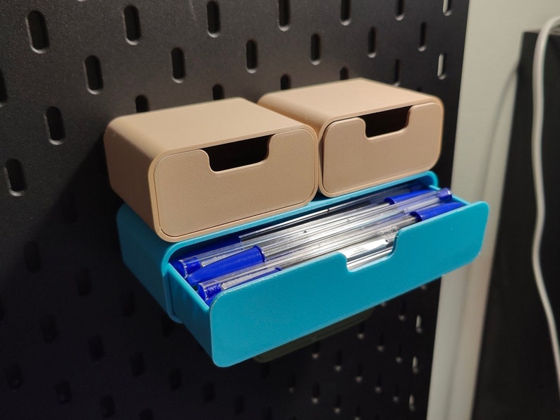 Puede incluir: Un conjunto de tres contenedores de almacenamiento en beige y turquesa. Los dos contenedores beige superiores tienen un dise&ntilde;o de caj&oacute;n. El contenedor turquesa inferior contiene bol&iacute;grafos azules. Montados en una pared perforada negra.