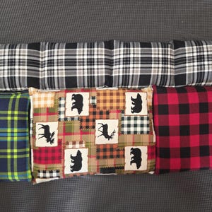 May include: A collection of decorative pillows with various plaid and patchwork designs. The pillows feature a mix of colors, including black, white, red, green, and brown. Some pillows have bear and moose silhouettes.