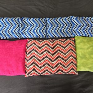 May include: An assortment of colorful fabric items, including rectangular and oblong shapes. The items feature various patterns, such as a blue and black chevron design, a red and black chevron design, a solid pink, and a solid green.