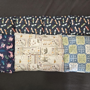 May include: An assortment of rectangular throw pillows with various patterns. The pillows feature designs such as dragonflies on a navy background, axolotls, a patterned beige design, and a blue and green geometric pattern.