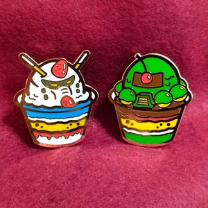 May include: Two enamel pins shaped like ice cream cups. One is white with red and blue accents and a strawberry on top. The other is green with brown accents and a cherry on top.