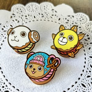 May include: Three enamel pins featuring characters from the anime One Piece. The pins are shaped like donuts and feature the characters Chopper, Luffy, and Zoro. The pins are gold-colored with colorful details.