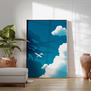 Peut inclure: Une affiche encadr&eacute;e repr&eacute;sente un avion traversant un ciel bleu avec des nuages blancs. L'&oelig;uvre est expos&eacute;e dans une pi&egrave;ce avec une grande plante en pot, un pouf gris et des poteries d&eacute;coratives.
