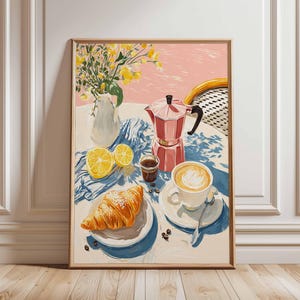 May include: A framed art print of a breakfast scene. A pink coffee maker, a cup of coffee, a croissant, and sliced lemons are arranged on a table. A vase of yellow flowers and a blue and white cloth complete the composition.