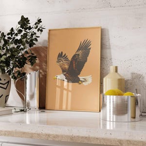 May include: A framed print featuring a detailed illustration of an eagle in flight. The eagle has brown and white feathers, a yellow beak, and is set against a tan background. The frame is gold-colored, and the print is displayed on a countertop.
