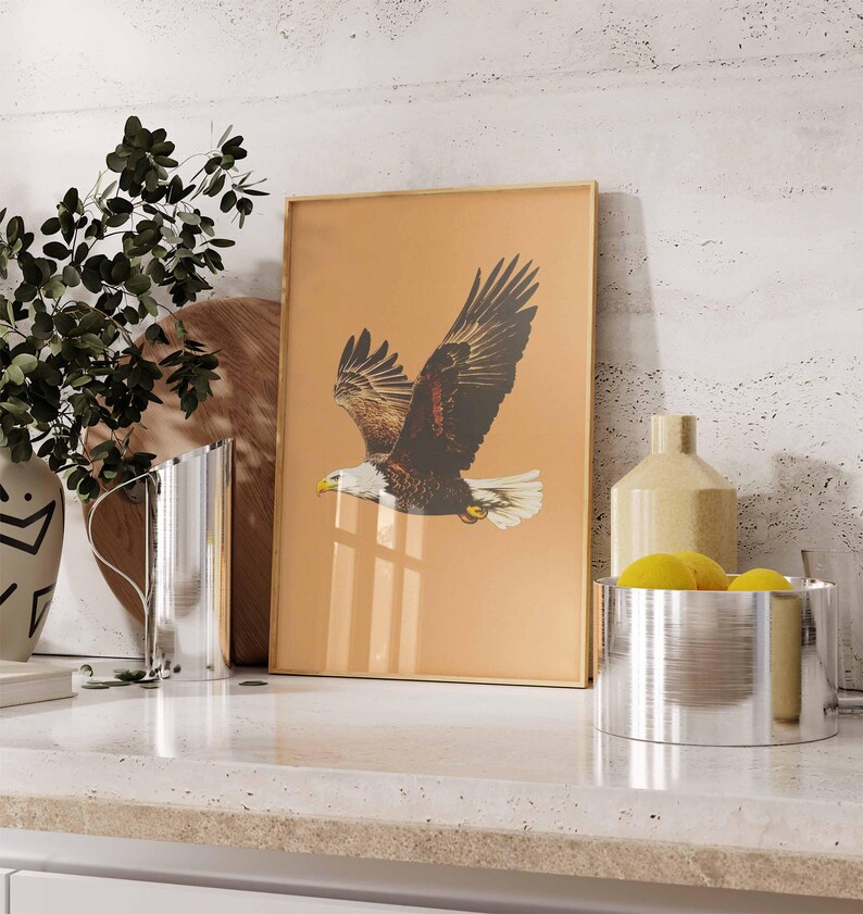 May include: A framed print featuring a detailed illustration of an eagle in flight. The eagle has brown and white feathers, a yellow beak, and is set against a tan background. The frame is gold-colored, and the print is displayed on a countertop.