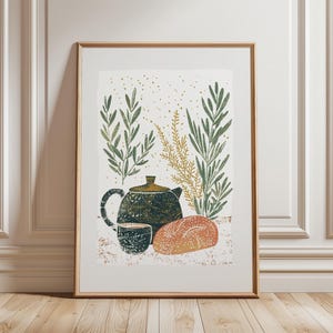 May include: A framed art print with a still life illustration. The artwork shows a dark green teapot, a matching cup, and a loaf of bread. Green and gold foliage surrounds the objects, with gold speckles in the background. The frame is a light brown colour.