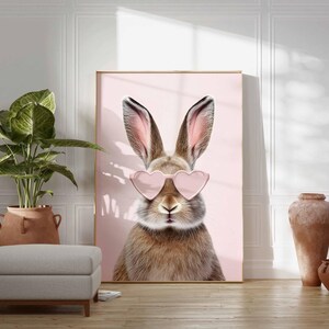 May include: A framed art print featuring a close-up of a brown rabbit wearing pink heart-shaped sunglasses against a pink background. The artwork is displayed in a room with a plant, ottoman, and decorative vases.