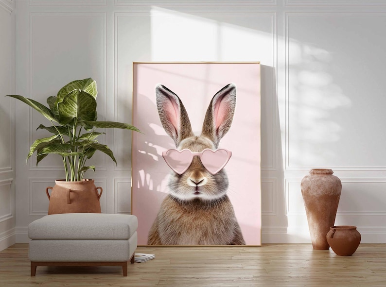 May include: A framed art print featuring a close-up of a brown rabbit wearing pink heart-shaped sunglasses against a pink background. The artwork is displayed in a room with a plant, ottoman, and decorative vases.
