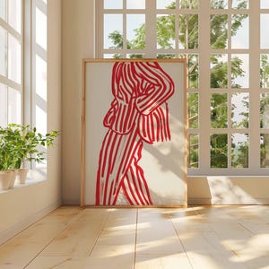 K&ouml;nnte beinhalten: Ein gerahmter Kunstdruck mit einer Figur in rot-wei&szlig; gestreifter Kleidung. Das Kunstwerk wird in einem Raum mit Tageslicht, Holzboden und einem Fenster mit Blick auf Gr&uuml;n pr&auml;sentiert. Der Rahmen ist hellbraun.