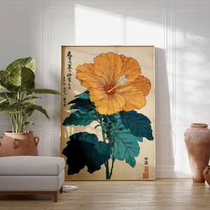 May include: A framed art print depicting a large orange hibiscus flower with green leaves on a beige backdrop. The print includes Japanese text and is displayed in a room with a plant, ottoman, and pottery.