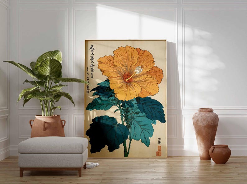 May include: A framed art print depicting a large orange hibiscus flower with green leaves on a beige backdrop. The print includes Japanese text and is displayed in a room with a plant, ottoman, and pottery.