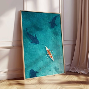 May include: A framed art print featuring an aerial view of a turquoise ocean with dark blue whale shark silhouettes. A small white boat with orange figures is in the center. The artwork is displayed on a wooden floor next to a white wall and a beige curtain.
